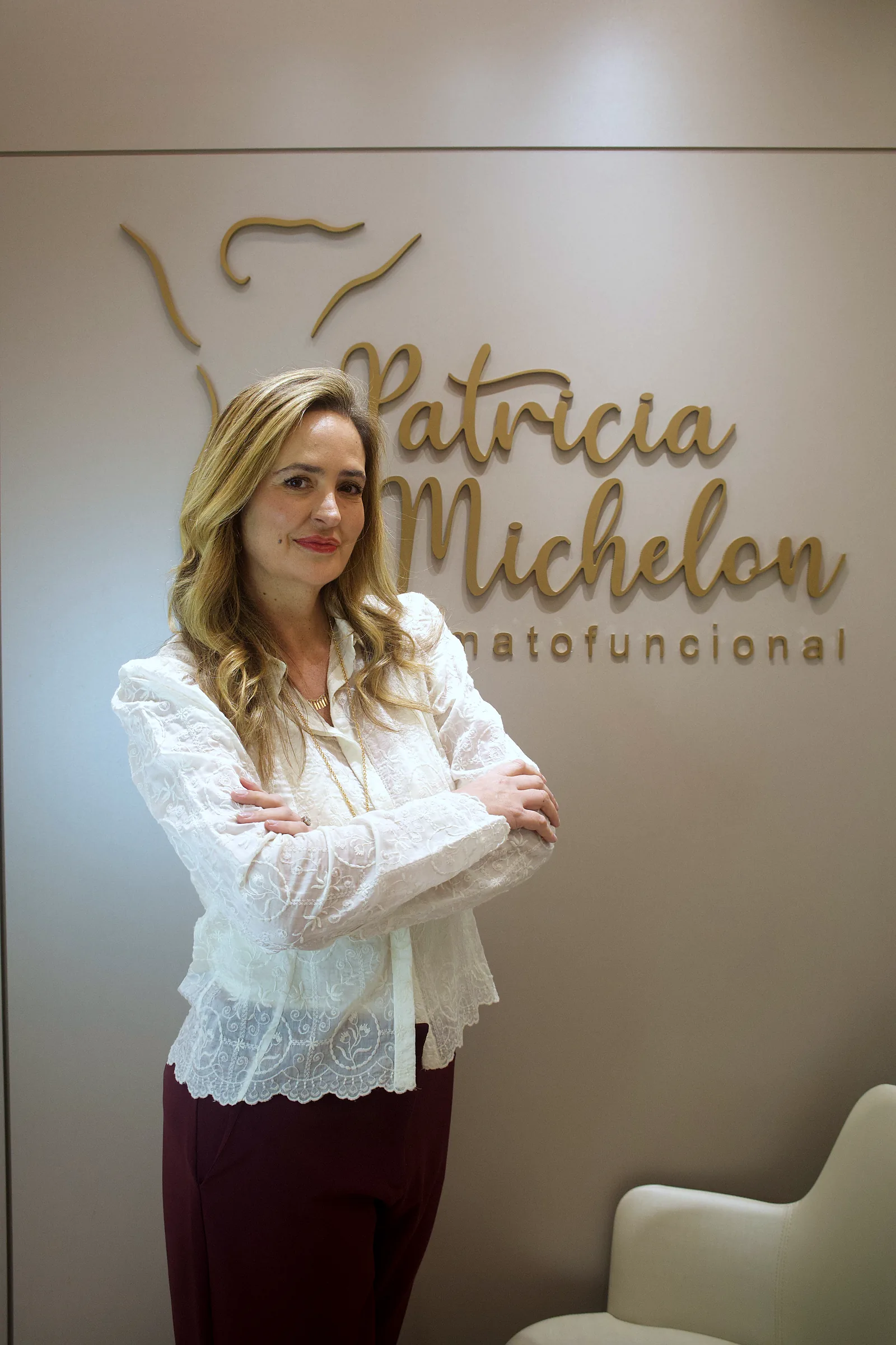 Patricia Michelon, fisioterapeuta dermatofuncional, de blazer de renda branca e braços cruzados, em frente ao logotipo dourado da clínica.