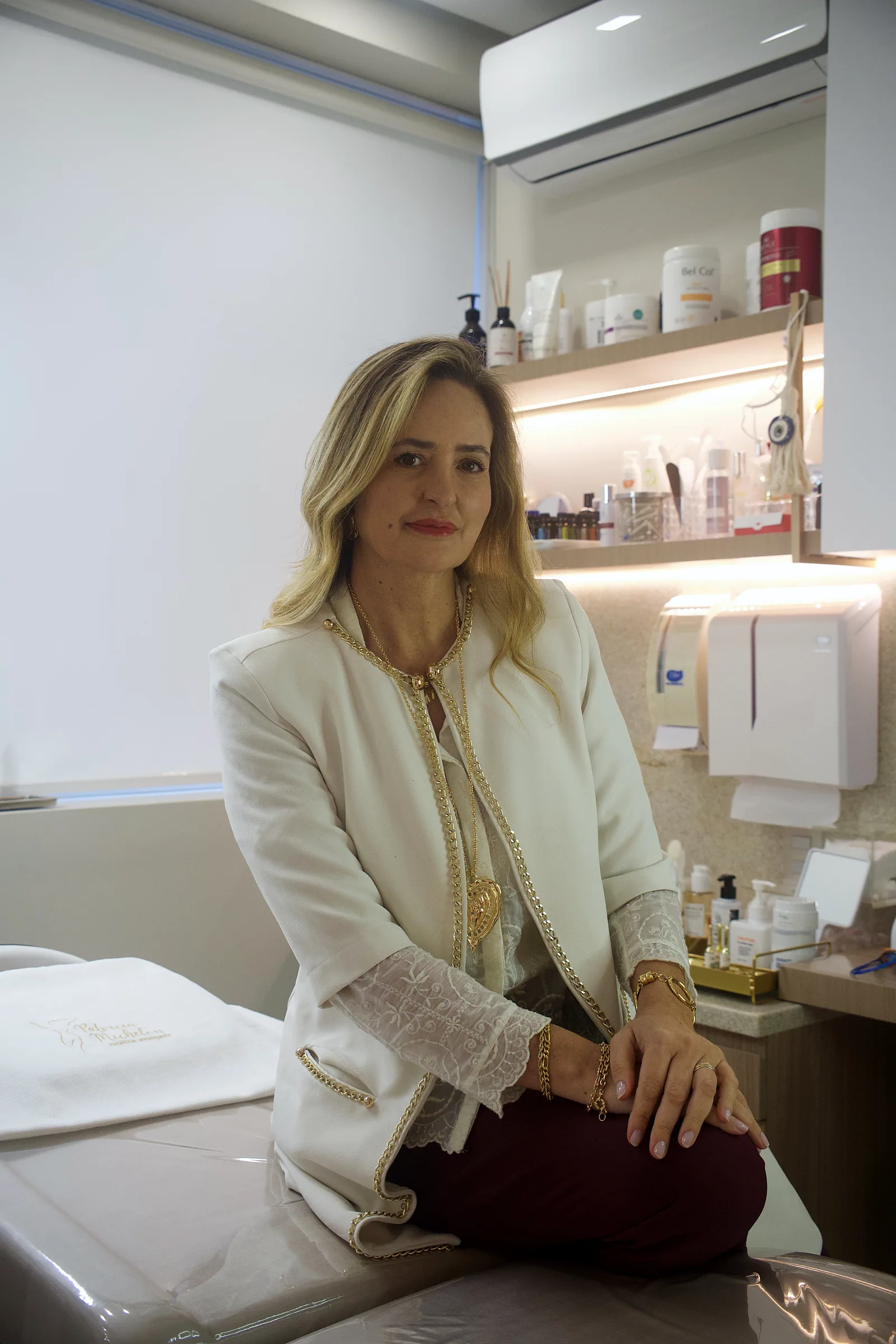 Patricia Michelon sentada na maca da sala de atendimento, de blazer branco.