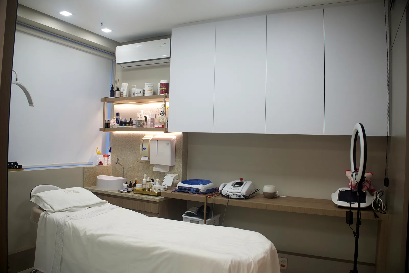 Sala de tratamento facial da clínica, com maca bege, lupa iluminada e equipamentos na bancada.